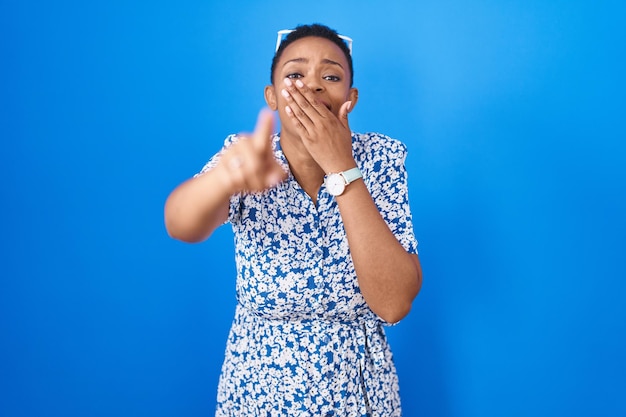 African american woman standing over blue background laughing at you, pointing finger to the camera with hand over mouth, shame expression