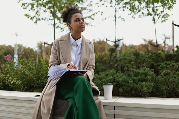 African american woman student making notes in a notebook while drinking coffee outside the office