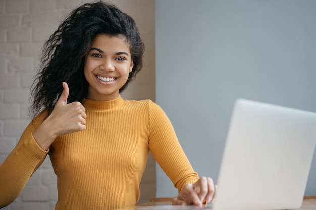 African American woman using laptop, showing thumb up, looking at camera and smiling