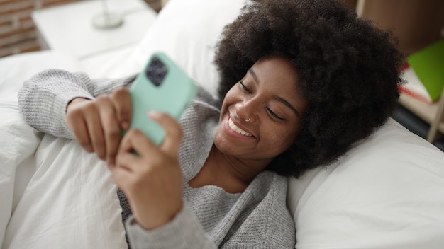 African american woman using smartphone lying on bed at bedroom