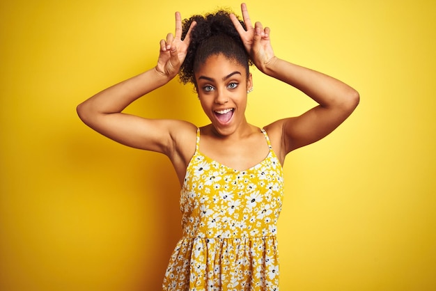 African american woman wearing casual floral dress standing over isolated yellow background Posing funny and crazy with fingers on head as bunny ears smiling cheerful