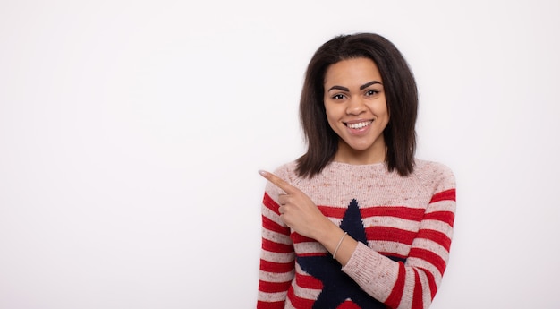 African american woman wearing casual pink striped sweater over isolated white wall smiling and looking at the camera pointing to the side.