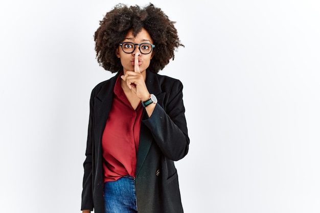 African american woman with afro hair wearing business jacket and glasses asking to be quiet with finger on lips silence and secret concept