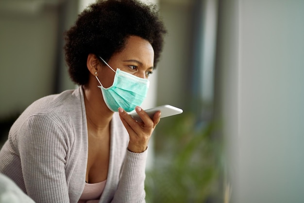 African American woman with face mask recording voice message on smart phone at home