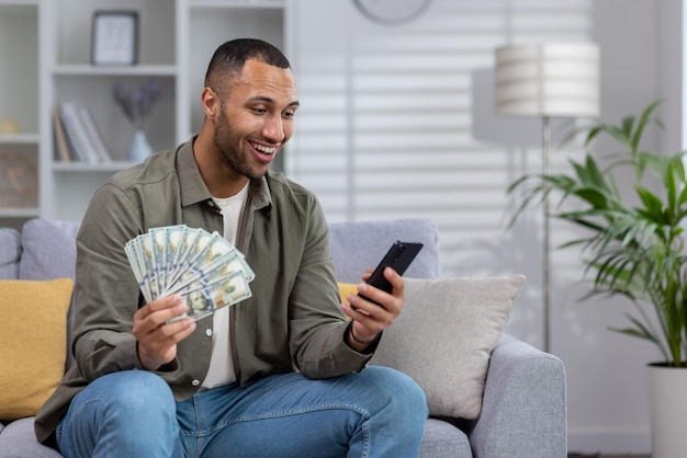 African american young man sitting on sofa at home and holding fan of cash money and dollars using