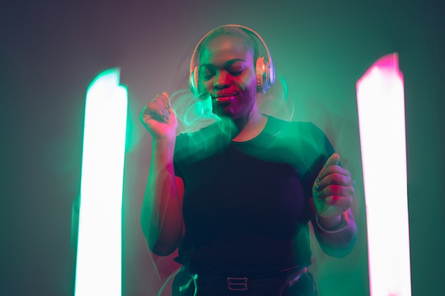 African-american young woman's portrait on gradient background. Beautiful female model dancing, singing in neon lights delightful. Concept of human emotions, facial expression, sales, ad, inclusion.