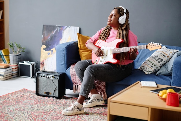 African american young woman sitting on sofa playing guitar and singing song