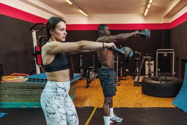 African athlete boy and Caucasian athlete girl in a gym doing shoulder biceps and pectoral exercises with kettlebells in a gym. Two multiracial bodybuilders