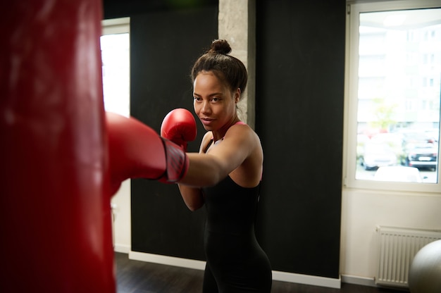 African athletic woman with perfect physique , boxer in red boxing gloves, trains hard, strikes a straight punch, hits a huge punching bag in a boxing gym. Martial art concept.