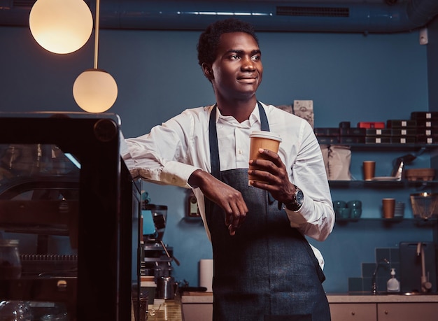 African barista relaxing after workday with coffee while leaning on counter at coffee shop.