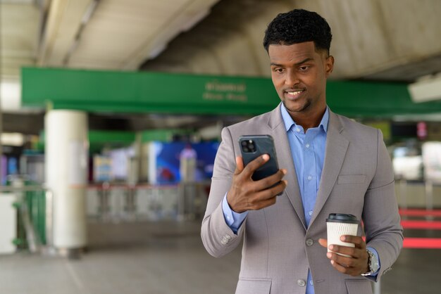 African businessman outdoors carrying cup of take away coffee while smiling and using mobile phone