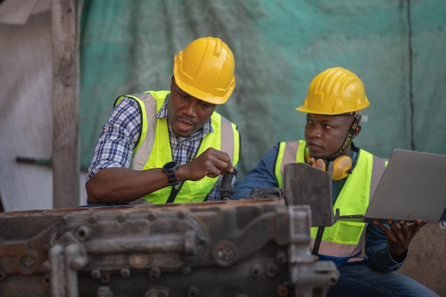 African Engineer Technician holding laptop for checking and inspecting on Heavy machine engine