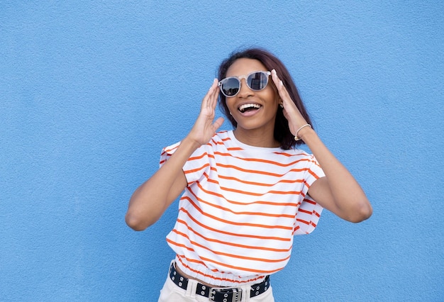 African ethnicity young woman in stylish sunglasses with curly hair tied up in high ponytail looking away while smiling broadly showing straight perfect teeth posing against blue wall background