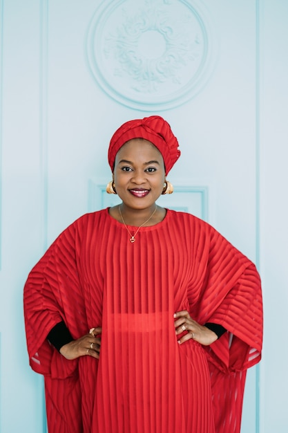 African fashion model in traditional african style with red stylish suit, head scarf, earrings and makeup on light blue door background in studio.