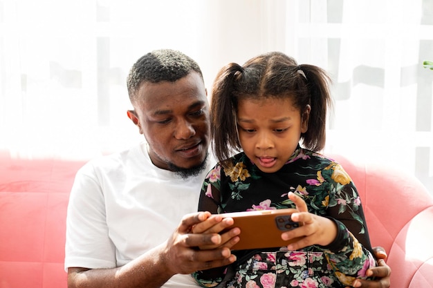 African father holding smart phone and teach his daughter in living room.Adult teaching and enjoying with sister on the sofa.