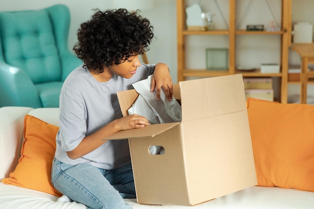 African girl unpacking delivery looking in box Happy woman opening carton box Female getting parcel looking at delivered goods items Satisfied client positive feedback quick express deliver