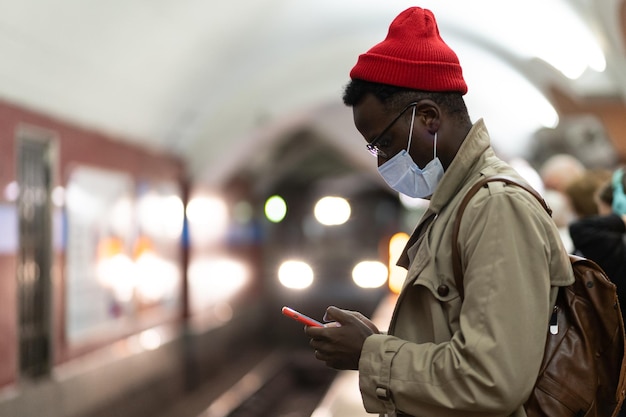 African man in face mask in subway station wait for train using smartphone scrolling social media