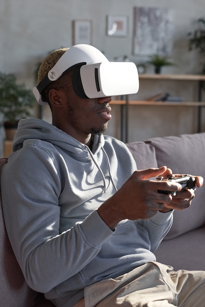 African man in glasses sitting on sofa and using joystick he playing video game at home