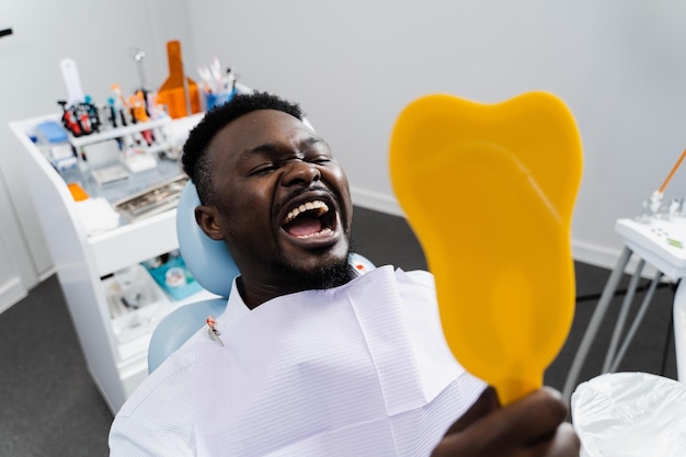 African man patient is looking in the mirror at his teeth after removing caries and filling teeth Consultation with dentist in dental clinic African american patient is visiting dentist