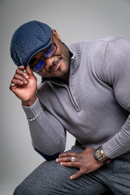 African man in tight shirt sitting and holding his cap Strong man with big biceps wearing a cap and glasses Muscular man in grey clothes in white background