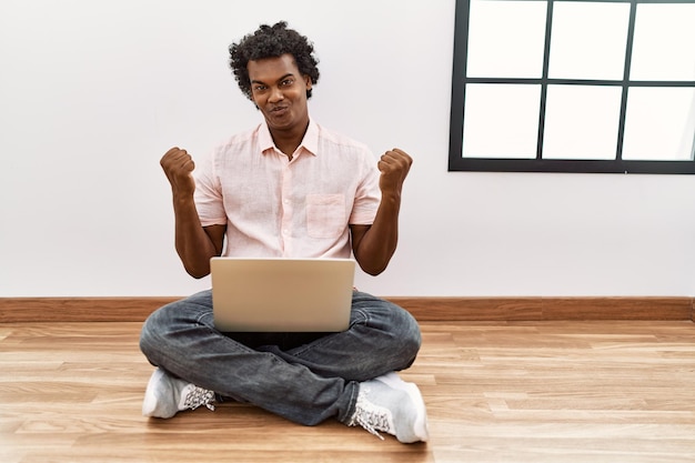 African man with curly hair using laptop sitting on the floor celebrating surprised and amazed for success with arms raised and open eyes. winner concept.