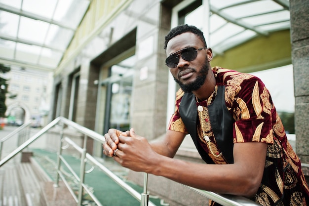 African stylish and handsome man in traditional outfit and sunglasses standing outdoor.