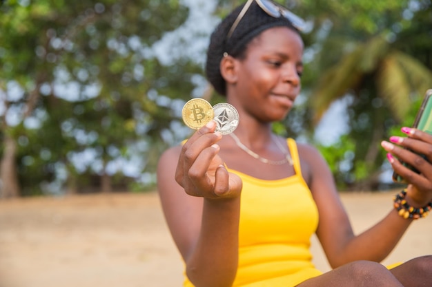 An african trader with Bitcoin and ethereum coins on the beach checks the market with her phone