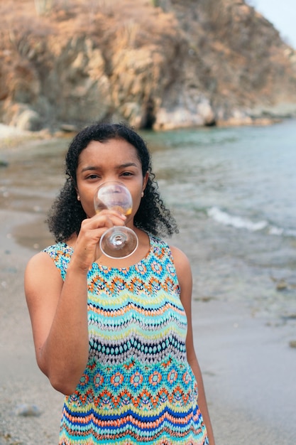 African woman drinking a glass of wine looking at the camera by the sea.