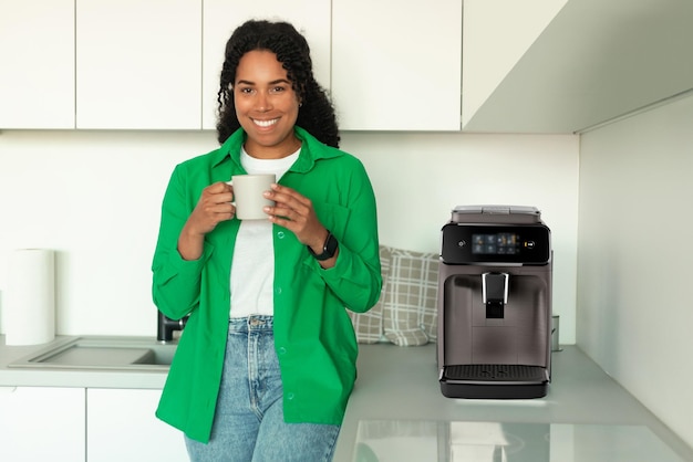 African Woman Holding Cup Standing Near Coffee Machine In Kitchen