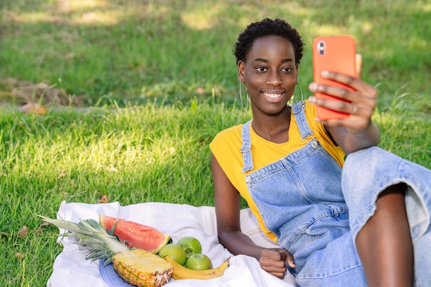 African woman taking a selfie with her mobile phone while having a picnic of fruits