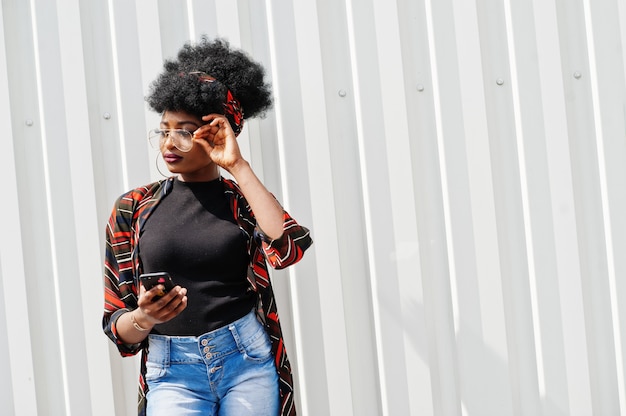 African woman with afro hair, in jeans shorts and eyeglasses posed against white steel wall with mobile phone in hand.