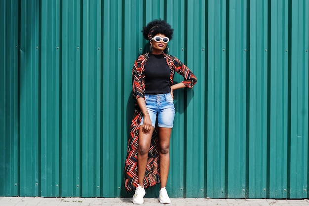 African woman with afro hair in jeans shorts and white sunglasses posed against green steel wall