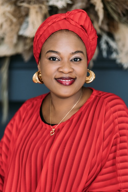 African woman with bright makeup and stylish jewerly, wearing red dress and ethnic headwrap, posing in studio