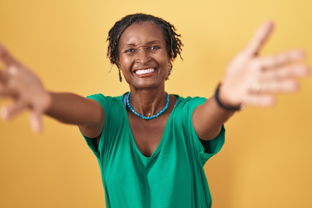 African woman with dreadlocks standing over yellow background looking at the camera smiling with open arms for hug. cheerful expression embracing happiness.