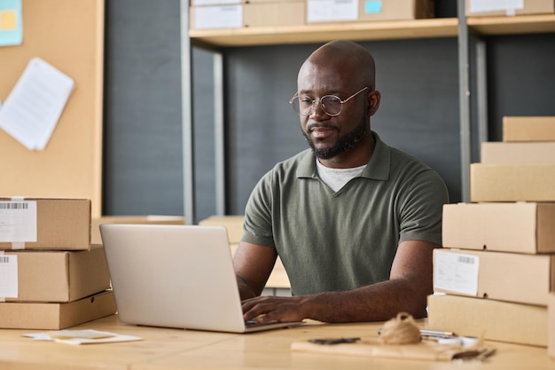 African worker typing on laptop making delivery online while sitting at table with stack of parcels