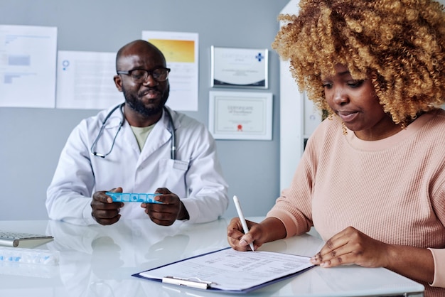 African young woman with curly hair signing insurance before treatment while sitting at doctors office