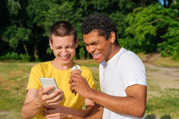 Africanamerican and caucasian friends eats ice cream and use smartphone outdoor