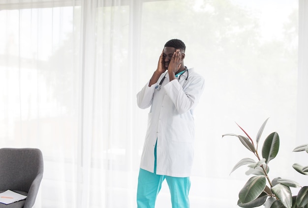 An AfricanAmerican doctor tired of hard work is standing near the window