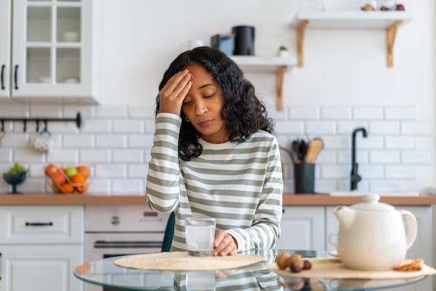 Africanamerican female dealing with headache dissolving pill in glass of water in kitchen