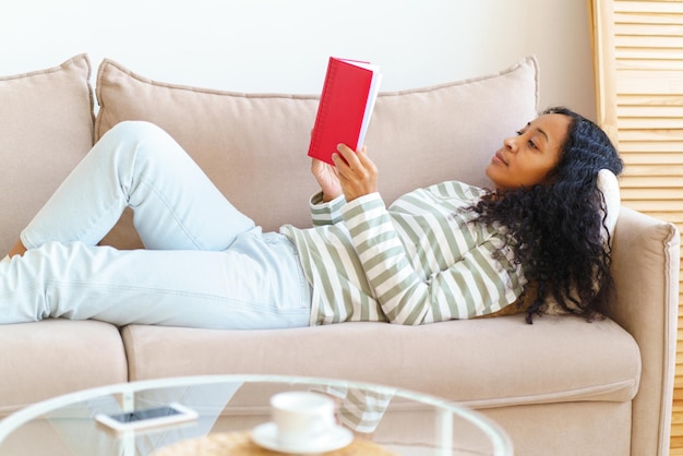 Africanamerican lying down on sofa and reading book in red cover slow paced lifestyle