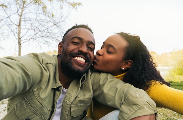 afro american couple taking a selfie kissing on the cheek sitting in a park