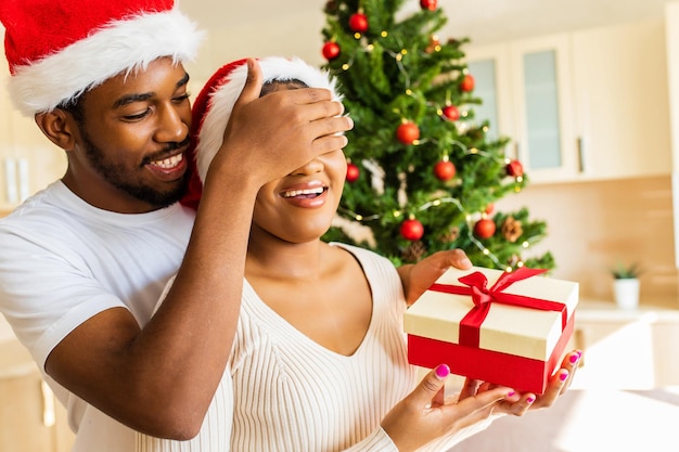 Afro american man giving gift box to his girlfriend in house near christmas tree