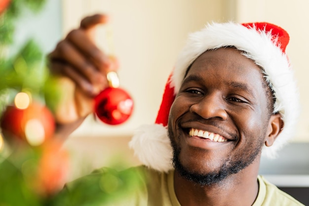 Afro american man in Santa hft decorate the christmas tree with a red ball at home