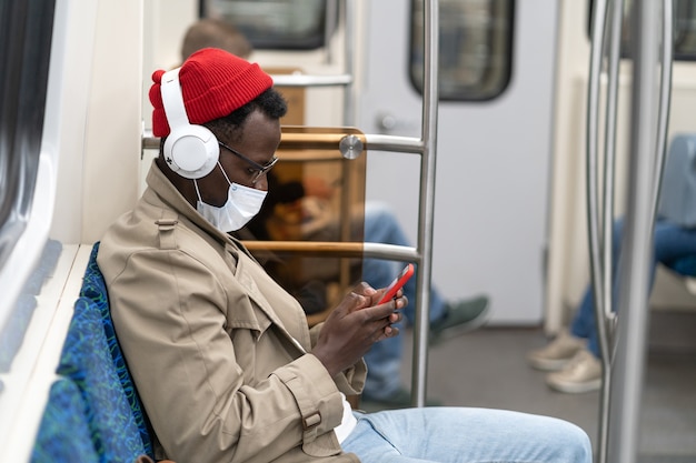 Afro-American man in subway train, wear face mask, using mobile phone, listens to music