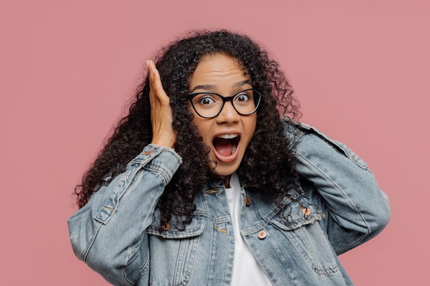 Afro American woman covers ears, yells loudly, ignores loud sound, keeps mouth widely opened