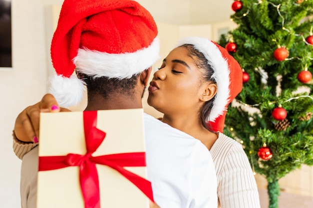 Afro american woman kissing on cheek of boyfriend for gift box in house near christmas tree