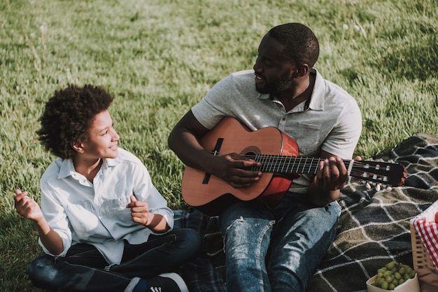 Afro Father Play on Guitar and Looking on Son in Picnic.