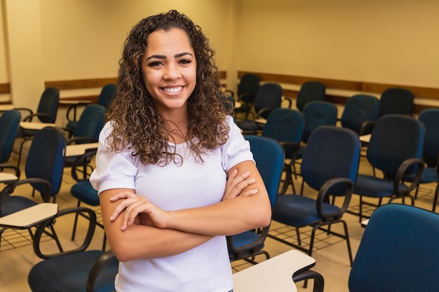 Afro student in classroom with arms crossed looking at camera smiling. Young student with the classroom in the background.