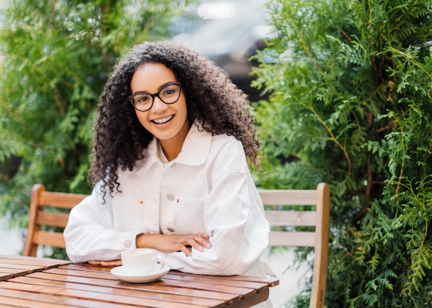 Afro woman in white clothes, drinks coffee in outdoor cafeteria