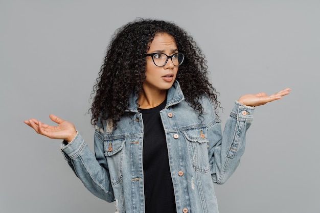 Afro woman with crisp hair, raises hands in bewilderment, looks aside